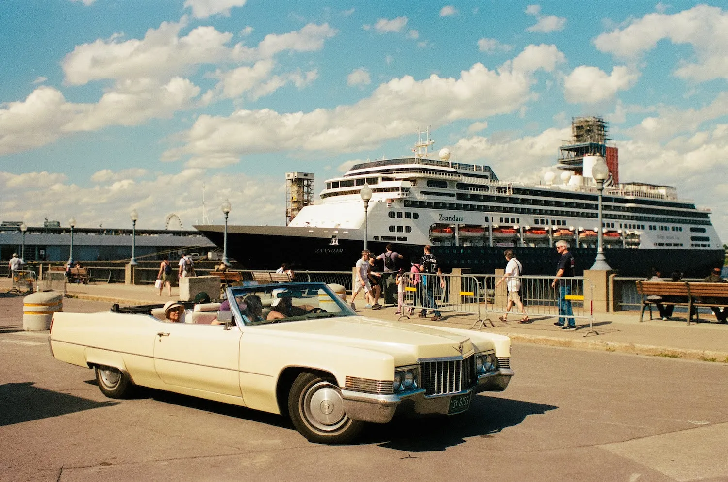 A classic vintage convertible car driving along a port harbor with a large cruise ship docked in the background.