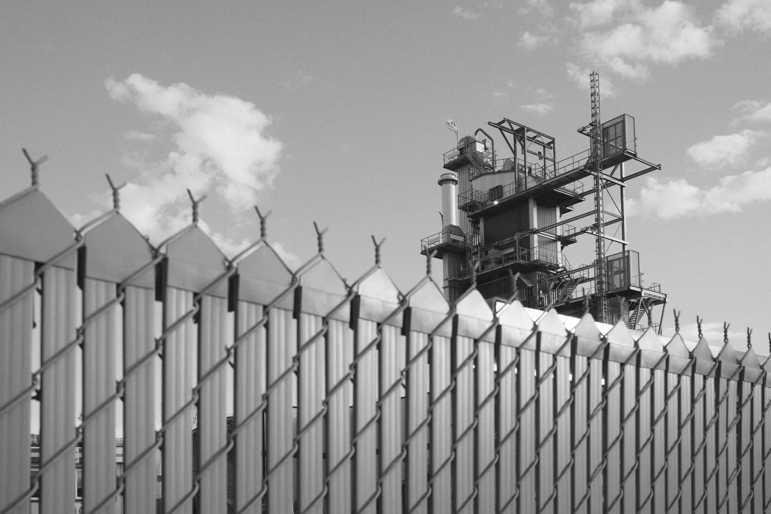 A black and white view of an industrial tower seen behind a fence.
