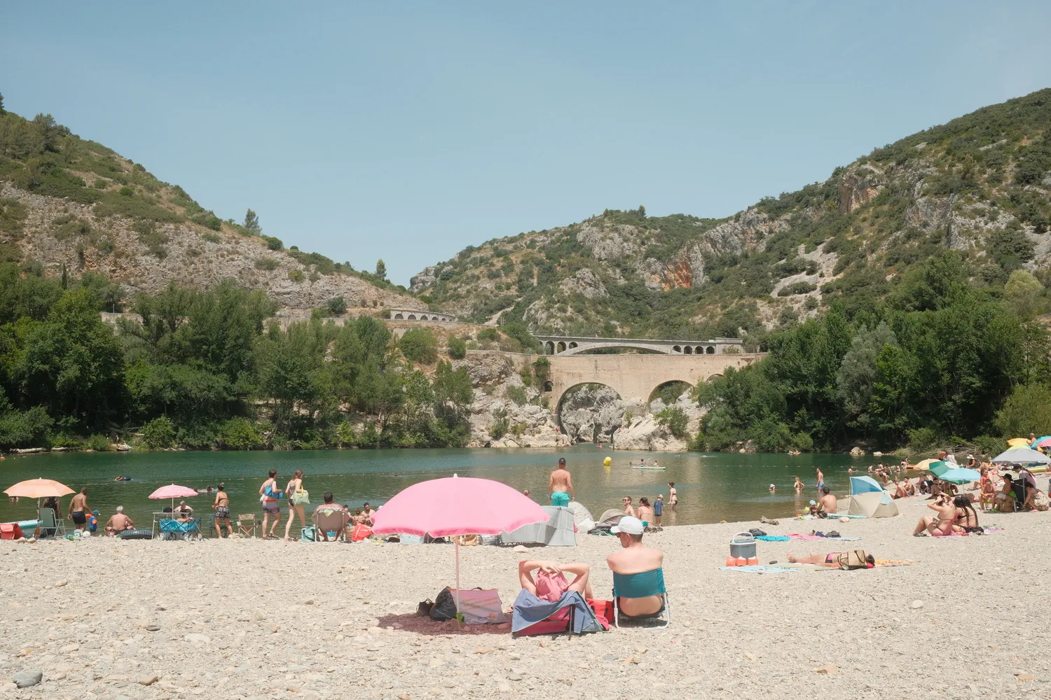 People relaxing under beach umbrellas on a pebbly riverbank with stone bridges in the background.