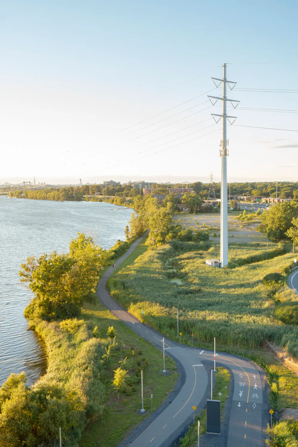 An elevated view of a winding bike path along a riverbank near a tall power line tower.