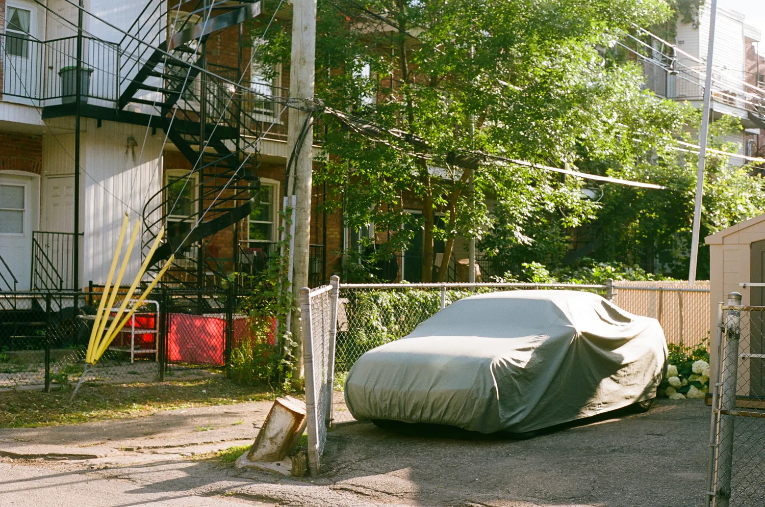 A car covered with a tarp in a residential back alley with outdoor spiral staircases.