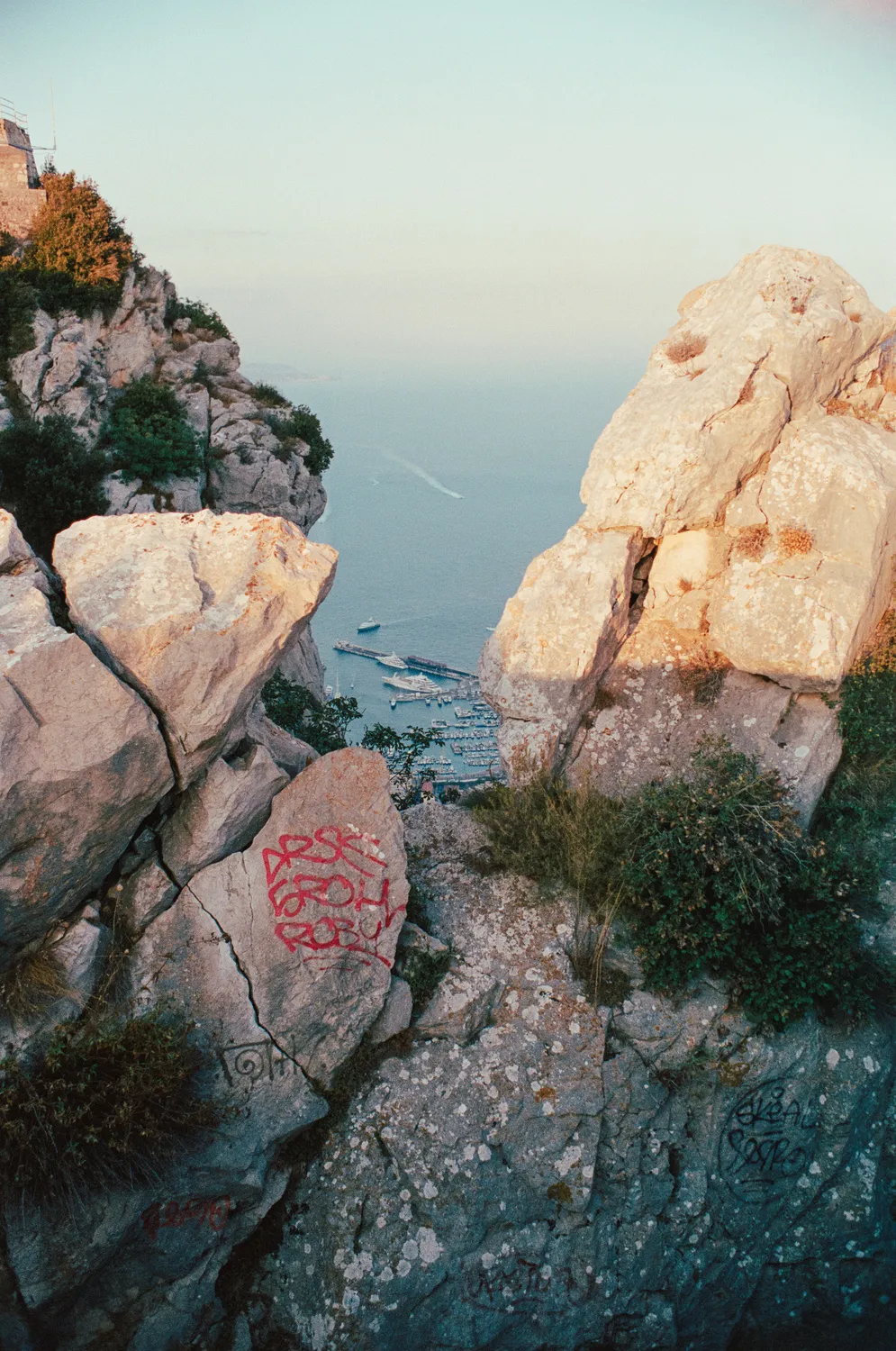 A view through rocky cliff formations looking down onto a harbor filled with yachts.